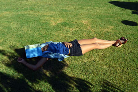 Tan young lady laying on the grass with a book over her faceの写真素材