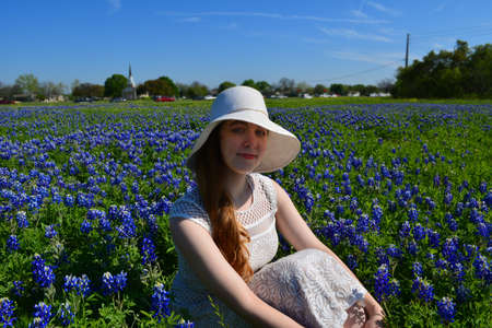 Beautiful girl in white bonnet sitting in a field of Texas Bluebonnet wildflowersの写真素材