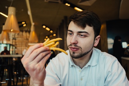 a young man with a beard sitting in a restaurant and holding hands french fries and going to eatの写真素材