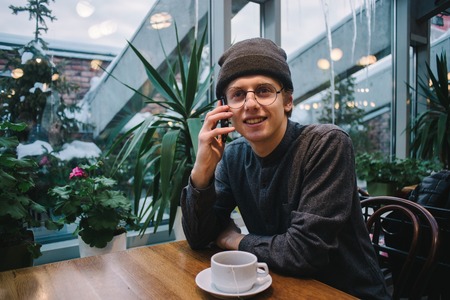 hipster smiling young man in glasses and a shirt, talking on the phone. coffee is on the table and the window greenhouseの写真素材