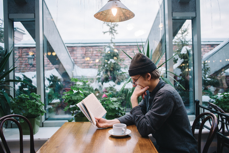 a young student in cap and shirt is reading a very interesting book and drinking coffee in a...