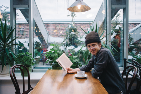smiling young hipster man spends his free time with interesting book and a hot drink in a nice cafeの写真素材