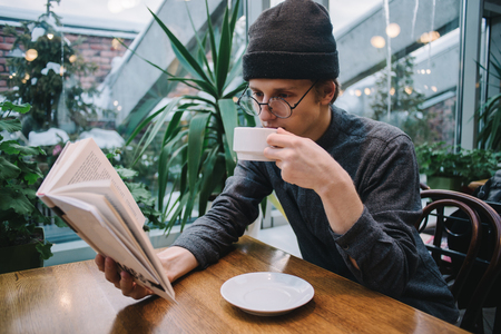 a young man in a cap and shirt reading a book and drinking coffee in a luxury restaurant with conservatoryの写真素材
