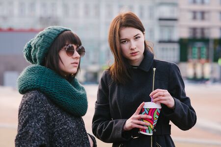 Portrait of two stylish girls friends are standing in the middle of the city. One girl is holding a coffee in another scarf and glassesの写真素材