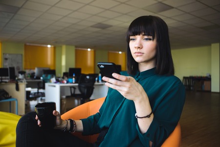 Portrait of smiling modern business woman in office using her mobile phone.Young handsome office worker in a modern office in background jobs. Business woman holding a cup of coffee. Break work.の写真素材