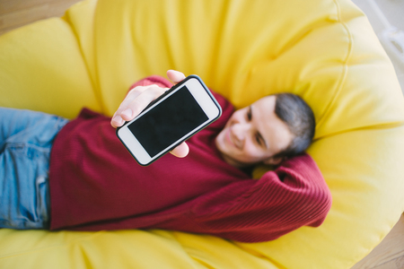 positive young man hipster sends phone camera while sitting in a yellow chair bagの写真素材