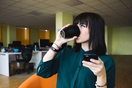 Portrait of a young, beautiful woman office worker that uses the mobile phone and drinking coffee from black cup. Break work. Modern officeの写真素材