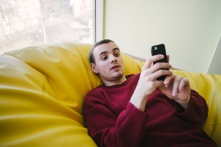Hipster young man lying in a large armchair bag and use the phone.の写真素材