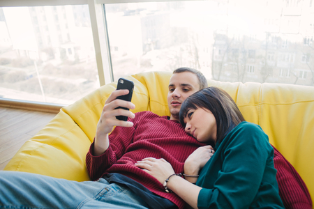 Young beautiful man and girl resting in a room with a modern interior and using a mobile phone. Bag chair in a modern apartment.の写真素材