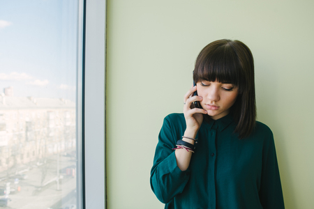 Young beautiful woman office worker talking on the phone at the window and looks down. Modern officeの写真素材