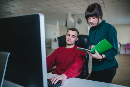 Young office workers. A young man sits at the computer in his chair and young business woman standing next to him and show something in a notebook.の写真素材