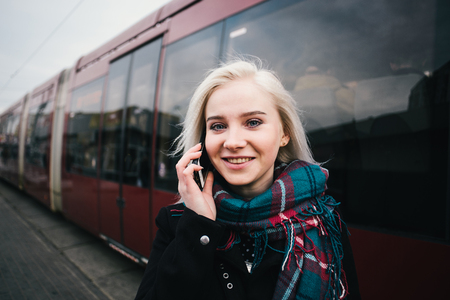 Street stylish portrait of a young and beautiful woman talking by phone on the background of modern train. She uses the phone at the station.の写真素材