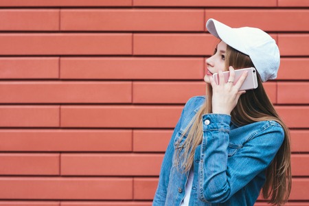 Stylish blonde girl in a denim jacket and cap talks by phone on a red background. A young woman uses a mobile phone hipsterの写真素材
