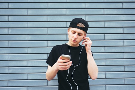 Beautiful young man in a cap and a black T-shirt gets listening to music on headphones with your phone. Student listening to music on a background of blue wallの写真素材