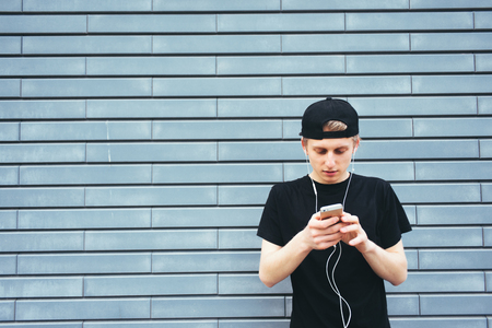 Student in cap and shirt listening to music on headphones typing on your phone. A young man talking on the Internet and listening to music on the background wallの写真素材