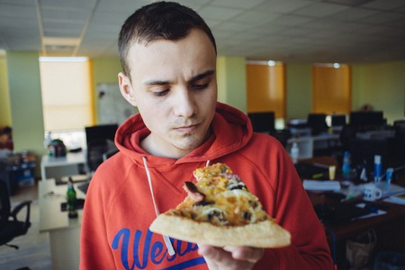 The young man eating delicious pizza against a background of office space. Fast food a break at workの写真素材