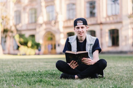 A smiling student reads a book in the countryside near the college campus. Teenager reading a book...