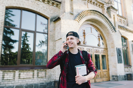 Portrait of a smiling student with books in his hands talking on the phone at the background of the university. A young man in a red shirt speaks against the background of the entrance to college.の写真素材