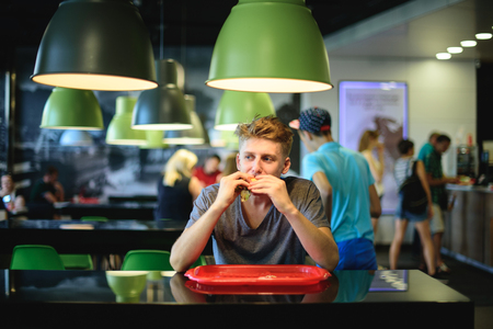 The teenager is sitting in a fast-food restaurant, eating a large, beautiful burger and looking away.の写真素材
