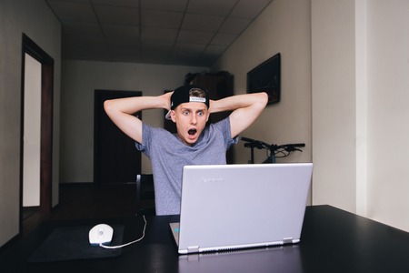 Amazed teenager with fear looks at the monitor of his computer while sitting at the table at home.の写真素材