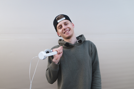 A positive young man stands near a light wall, holds headphones and smiles.の写真素材