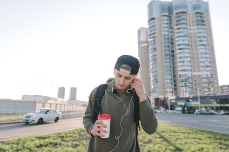 A handsome man with a glass of hot drink in his hands listens to music and corrects headphones in his ears against the backdrop of urban scenery. Life stylesの写真素材