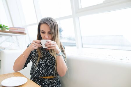 Beautiful young woman in a cafe holding a cup of tea, seen through the window with buildings and...