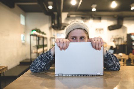A funny young man hiding behind a laptop. Cheerful hipsters work at the computer in a cozy cafe. Look away.の写真素材