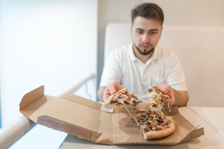 A man sits at the table near the box and holds in his hands a few pieces of pizza. The man eats a...