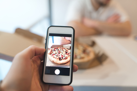 A man makes a photo of a box of pizza on his cell phone. Photo of fastfood on a smartphone.の写真素材