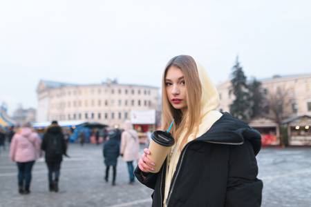 A beautiful girl stands in the background of a citys winter street with a cup of coffee in her hands and looks at the camera. The girl is warm with a hot drinkの写真素材