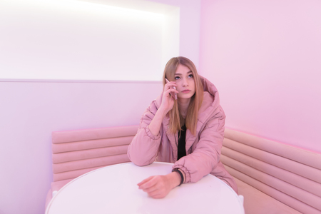 Cute girl in pink dress sitting at the table in a room with pink interior, talking on the phone and looking up.の写真素材