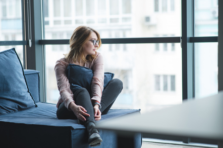 Beautiful girl sitting on a sofa near the window and looking away. Girl office worker resting on the sofa in the officeの写真素材