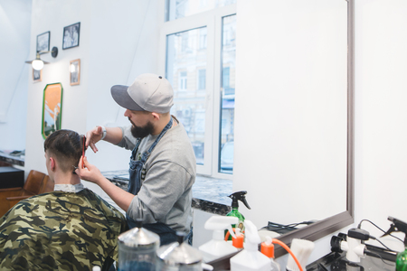 Barber with a beard cuts a young man with scissors in a men's beauty salon. Men's haircuts in barbershop.の写真素材