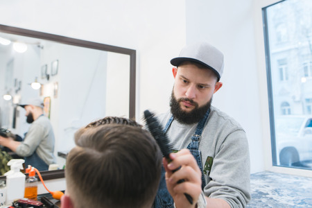 Barber with a beard creates a stylish hairstyle for a young man in a barbershop. A serious...