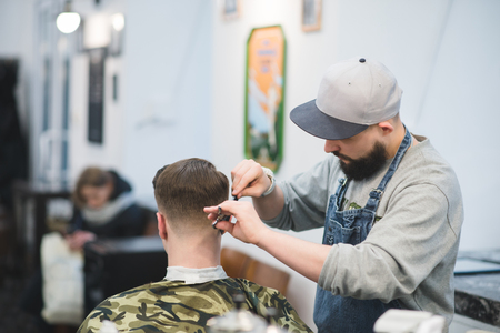 Haircut in barbershop. The male hairdresser with a beard makes a hairstyle to the client. Barber cuts a young man in a man's hairdressing salon.の写真素材