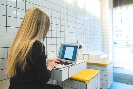 A student wears glasses at the table in a cafe with a stylish interior. The girl is sitting at the table and looks at the notebook monitor. Work on a computer in a public place.の写真素材