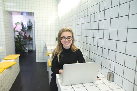 A smiling student with a laptop in a cafe with a light interior. . Positive girl sitting in a cafe at a table near a laptop and smilingの写真素材