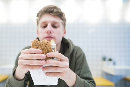 Hungry young man looks at a hamburger in his hands. The student eats fast food in a light restaurant with a stylish interior.の写真素材