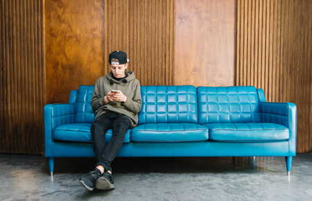 Young man sitting on a blue couch and using a smartphone. The student is sitting on the couch and writes a message on the swarthy. On the background of a wooden wall.の写真素材