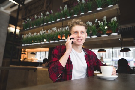 A student talks by phone in the cafe and smiles. A young man drinks coffee and talks over the phoneの写真素材