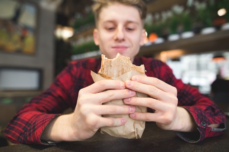 A student eats a sandwich in a cozy cafe. A young man with a happy look looks at the panini sandwich. in their hands. Focus on the sandwichの写真素材