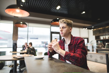 A student eats a sandwich in a cozy cafe and looks thoughtfully in the window. A beautiful young man alone dine in a restaurantの写真素材