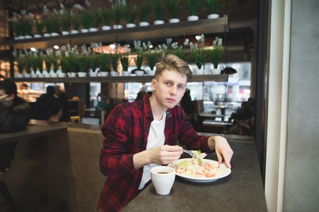 A young student in a red shirt eats a salad in a cozy cafe and looks at the camera. A student eats in a cafe.の写真素材