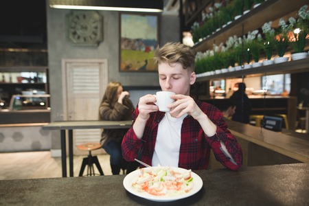 Young man with pleasure drinking coffee while eating in a cozy cafe. A student drinks coffee and eats a salad. Lunch Breakの写真素材