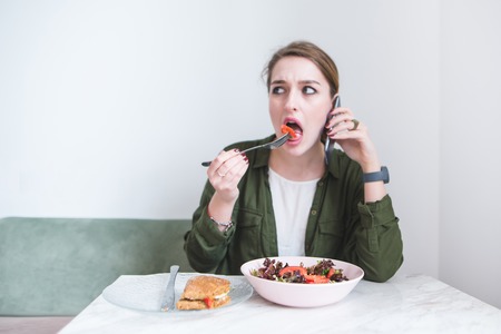 Business woman eating salad and talking on the phone in a restaurant on the background of a light wall. Lunch break in the cafe.の写真素材