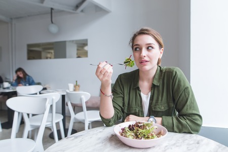 Beautiful young woman sitting in a cozy light restaurant with a fork in her hands, eating salad and looking away. A good meal for lunch at the restaurant.の写真素材