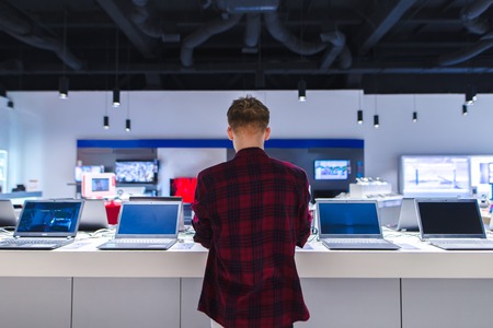 A young man stands behind his laptop at the electronics store. A young man chooses a laptop in the store.の写真素材