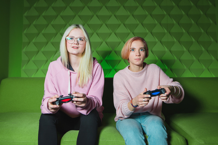 Two friends are focused on the monitor while playing console games. Blondes with gamepads in their hands sit on a sofa that is on a green background.の写真素材