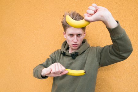 A funny teen is playing bananas as a weapon against an orange wall. Young man with bananas in his hands on a colored background.の写真素材
